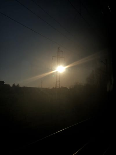 The image displays an outdoor scene in Dettwiller, France, characterized by strong backlighting during either sunrise or sunset. A prominent lattice-style electrical transmission tower stands centrally in the mid-ground, largely silhouetted, with a brilliant sun positioned directly behind its upper structure. Intense solar glare and a horizontal lens flare extend across the image from this light source. A smaller, similar transmission tower is faintly visible further in the background to the left. The horizon consists of a dark, indistinct landmass, possibly a hill, topped with sparse, silhouetted vegetation. A dark building silhouette is present on the far left. Several dark, thin power lines traverse the upper sky. The foreground is heavily shadowed, revealing two faint, parallel diagonal lines consistent with railway tracks. No individuals are present. The atmosphere appears hazy or diffused.