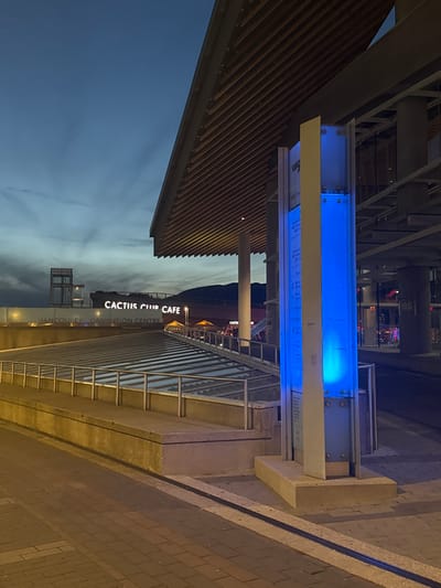 This image captures an exterior view of the Vancouver Convention Centre at dusk. A large, modern building with a distinctive wooden slatted roof overhang dominates the upper right portion of the frame. To the left, the text "CACTUS CLUB CAFE" and "VANCOUVER CONVENTION CENTRE" is illuminated against the darkening sky, identifying the establishment and its location. A pathway paved with interlocking bricks leads towards the foreground. A concrete structure with a metal railing runs along the left side of the pathway, suggesting a tiered entrance or walkway. A prominent blue illuminated pillar stands on the right side of the pathway, likely a sign or informational post. In the background, streetlights and building interiors are visible, indicating an urban environment. The sky is a gradient of deep blue and grey, with faint cloud patterns. No people are clearly visible in the frame.