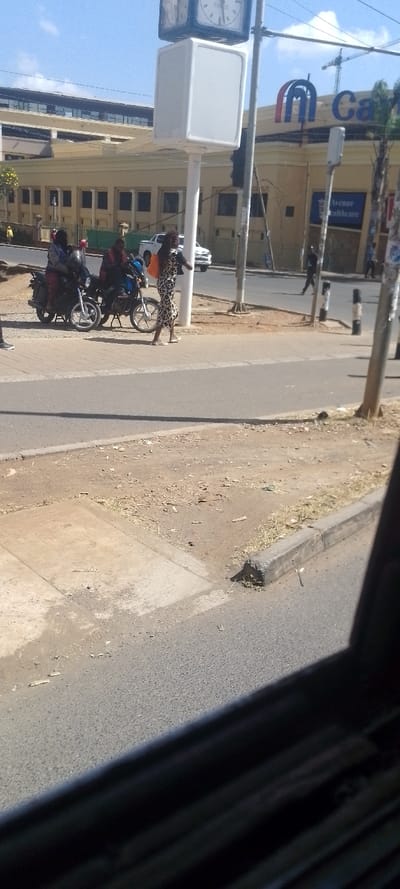 The image displays an urban street scene in Karen, Ngong Ward, Kenya, under clear, sunny conditions. The lower right foreground is partially obscured by a dark, diagonal structure, consistent with a vehicle's interior. The mid-ground features an asphalt road bordered by a paved sidewalk and an unpaved dirt shoulder with sparse dry vegetation.
On the paved area adjacent to the road, two individuals are seated on motorcycles. One wears a light blue jacket over a dark garment, and the other wears a red top. A woman in a patterned dress, carrying an orange bag, walks rightward on the sidewalk. A tall white pole with a square clock at the top and a blank white rectangular sign below it stands near the road. Adjacent to it are traffic lights and multiple dark street poles, some marked with vertical black and white stripes (bollards). A person is visible walking across the street in the mid-ground right.
In the background, a prominent, light-colored building with a rounded facade is visible. It features a large "Can" logo (part of "Karen") and an "Avenue Healthcare" sign. Behind this building, another structure with a darker roofline is present. A white pickup truck is parked on the far side of the street, and a construction crane is visible in the distant background against a clear blue sky.