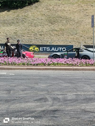 Two individuals, one wearing a black t-shirt and sunglasses, and the other in dark work overalls and a baseball cap, stand beside a red sports car. Behind them, a banner displays "ETS.AUTO" and a yellow logo of a car. A grey sports car with its door open is parked further down, partially obscured by pink flowers. The scene appears to be outdoors, with a grassy slope in the background and a paved road in the foreground. The image also contains a timestamp in the lower right corner: "27 июня 2026 г. в 14:03". The reference to Hrodna, Belarus is not directly verifiable from the visual information alone, but the timestamp format suggests a Cyrillic origin.