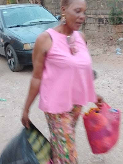 The image displays a female subject walking on an unpaved dirt and gravel path in Orie Uratta, Nigeria. The woman is in motion, wearing a pink sleeveless top, multi-colored patterned trousers, large gold-toned rectangular earrings, and a pendant. She carries a dark-colored bag in her left hand and a red plastic container in her right hand. In the background, a dark-colored sedan with five-spoke wheels is parked on the left, while a weathered concrete block wall is visible on the right.