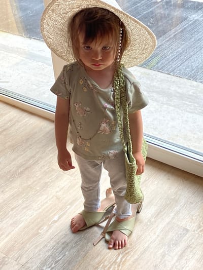 A young child is standing on a light-colored wooden floor, indoors, in front of a large window. The child is wearing a wide-brimmed straw hat with decorative pearls, a light green t-shirt with a bunny print, and gray pants. The child has a green crocheted bag slung over their shoulder and is standing in a pair of light green heeled sandals, one of which is placed on top of the other. The child's expression is serious, and they are looking downwards. The lighting suggests it is daytime. The background shows the window with reflections and possibly an outdoor scene, but it is blurred and indistinct. There is no information in the image to suggest this location is in the city of Tyumen, Russia.