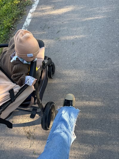 In the city of Мещёры, Russia, an outdoor scene depicts a close-up perspective of a person walking alongside a baby in a stroller. The pavement, marked by a white line on the left side, suggests a path or sidewalk. Sunlight casts dappled shadows across the asphalt. The baby, wearing a tan beanie with "happy days" embroidered on it, is nestled in a stroller with beige and brown fabric. The baby's hand, adorned with a patterned cloth, rests on the stroller's handle. The walker's legs, clad in ripped denim jeans, and a sneaker are visible in the lower right corner, indicating forward motion. The environment is characterized by a grassy verge on the left, suggesting a park or a tree-lined street.