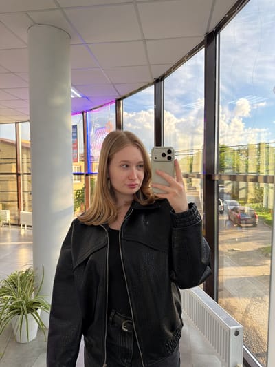 A fair-skinned young woman with blonde hair is taking a selfie with her iPhone inside a building in Rossiyskiy, Russia. She is wearing a black leather jacket. The building has large windows showing reflections of other buildings, the sky, and clouds. A white radiator is visible near the window. A large, white cylindrical support column is in the foreground. A potted plant is in the lower right corner. A store with neon lighting can be seen through the windows.