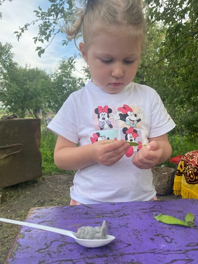 A young child with blonde pigtails, wearing a white t-shirt featuring a collage of Mickey Mouse characters, is engaged in an outdoor activity. The child is holding a green pea pod, examining it with a focused expression. A single pea pod is also visible resting on a purple wooden surface, next to a white plastic ladle containing a small, gray toy animal with blue eyes and an orange beak.

The setting appears to be outdoors, with lush green trees and foliage in the background, and a blue sky visible above. The ground is a mix of dirt and grass. A decorative fabric with gold and red patterns is partially visible in the upper right corner, suggesting a festive or decorative element in the environment. The lighting suggests it is daytime, possibly late afternoon or early evening given the soft light. The child's engagement with the pea pod and toy indicates a moment of play or exploration. The overall mood seems peaceful and focused on the child's interaction with nature and toys. There is no visible text in the image.