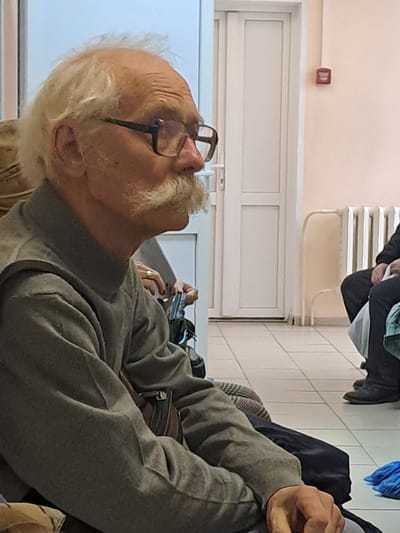 An elderly man with white hair and a mustache, wearing glasses and a gray sweater, is seated in what appears to be a waiting room. He looks upwards and to the left. Behind him, there is a closed white door with a red alarm pull station mounted on the wall above it. Further back, there is a white radiator and tiled wall. The location is likely Elektrostal, Russia. Another person is seated nearby, holding what appears to be a cell phone or small electronic device.
