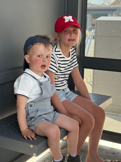 Two young children are seated on a gray, modern metal structure, possibly a bench or staircase. The girl, wearing a red baseball cap with a white letter 'M', a black and white striped shirt, and denim shorts, is positioned behind the boy. The boy, with a blue baseball cap, is dressed in a blue and white striped overall shorts set over a white shirt. He is lying on the metal structure. The setting is outdoors, with a perforated metal screen visible in the background, suggesting a building or a structure related to public transport or a recreational area. The location is Bredene, Belgium.