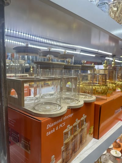 The image shows a display shelf in a retail setting, likely a shop in Kara-Suu, Kyrgyzstan. The shelf is illuminated by white LED strip lights. Prominently featured are a set of glass storage tanks with bamboo lids and crystal knobs, displayed on a wooden stand. These tanks appear to be part of a "Diamond storage tank 6 PCS" set. To the right of the glass tanks, there are several ornate golden decorative bowls or vases. The background shows shelves filled with more glassware and merchandise, some of which are out of focus. The foreground includes an orange cardboard box with product images and text, also related to the storage tanks. The overall impression is of a well-lit, organized retail display of kitchenware or decorative home goods.