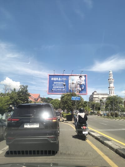 This media file captures a street scene in Banda Aceh, Indonesia, on a bright, sunny day with a few wispy clouds in a clear blue sky. The foreground shows the rear of a black SUV with the license plate "BL 1271 EO". To the right of the SUV, a person on a scooter, wearing a black jacket and a hijab, is also heading down the road.
Dominating the middle ground is a large billboard advertising "AQUA" mineral water. The text on the billboard reads, "AQUA 100% MURNI DINGIN ALAMINYA ADEMIN KELUARGA," suggesting the water is pure, cold, natural, and refreshing for the family. The billboard features images of a man and two children. To the right of the billboard, a tall, white minaret of a mosque stands prominently against the sky, indicating a likely Islamic cultural context. Further in the background, modern buildings and lush green trees line the street.
A blue sign with white text on the left side of the road reads "Kawasan Tertib Lalu Lintas," which translates to "Orderly Traffic Area." The road is paved with yellow lines marking lanes and the curb. The overall scene suggests a busy urban environment during daylight hours, with traffic flowing under a clear sky. The presence of the mosque minaret and the Arabic script on the HALAL certification on the billboard provide specific location cues.