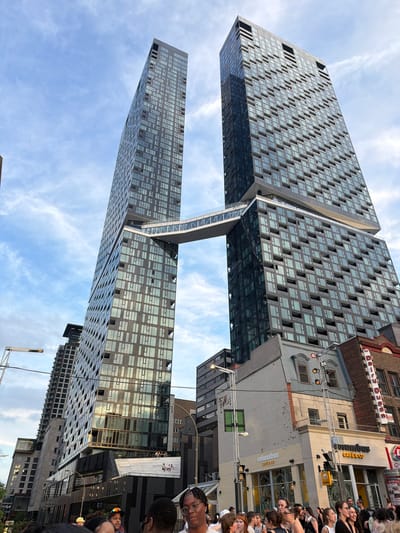 The image shows the unique architecture of the "Evo Montreal" residential complex, characterized by two towers connected by a skybridge. The towers are tall, modern, and clad in glass. In the foreground, a crowd of people is gathered, appearing to be at street level, looking up at the buildings. Several of them are looking directly at the camera. The sky is partly cloudy, with patches of blue visible. The scene suggests an urban environment, likely in Montreal, Canada, given the distinct architectural style often found in the city.