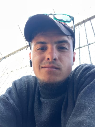 A medium close-up, upward-angle self-portrait of a man, likely in his late teens or early twenties, wearing a dark baseball cap with green-rimmed glasses perched on top, and a dark gray long-sleeved shirt or sweater. He has short dark hair, dark eyebrows, and a light beard. He is looking directly at the camera.

Behind the man, a wire mesh fence or trellis is visible against a bright, overcast sky.

Without further information, pinpointing the precise location in Yambol, Bulgaria, is impossible.