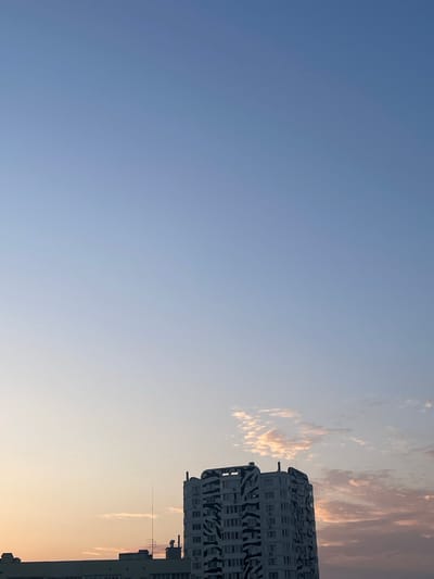 The image captures a high-rise residential building with a distinctive black and white patterned facade, located in the city of Brovary, Ukraine. The building is a prominent subject, positioned in the lower right quadrant of the frame. A lighter colored, lower-rise building is visible to the left of the patterned building. The sky above transitions from a clear, pale blue at the top to softer hues of pink and orange near the horizon, suggesting it is either dawn or dusk. Scattered, wispy clouds are present in the sky, tinged with the colors of the sunset or sunrise. The overall scene is urban, depicting a residential area with architecture typical of a post-Soviet country. No people are visible in the image.