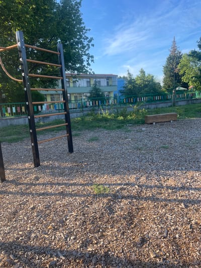 An outdoor scene in the city of Chirpan, Bulgaria, shows a playground area covered with wood chips. In the foreground, a pull-up bar structure with black metal uprights and wooden bars is visible. Behind the playground equipment, a green metal fence separates the area from a building with a light green and blue facade. Trees and greenery are present throughout the scene, with a clear blue sky overhead. A wooden bench is situated to the right of the playground equipment. The lighting suggests it is daytime, with shadows cast by the structures. There are no people or animals visible in the image.