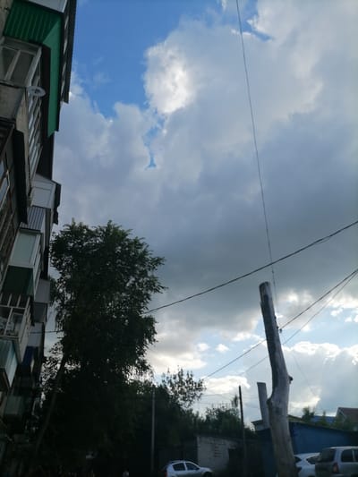 The image captures a view from an urban setting, likely Ulyanovsk, Russia, looking upwards and outwards. A multi-story building with balconies and green awning details is visible on the left, dominating the lower left quadrant of the frame. A large, leafy tree stands tall in the mid-ground, partially obscuring the building and extending towards the sky. Above, a dramatic sky unfolds with a mix of bright blue patches and thick, grey clouds, suggesting an overcast or impending weather change. Power lines are strung across the upper portion of the image, attached to a weathered wooden post that stands prominently in the right half of the frame. In the background, hints of other structures and trees are discernible, along with a glimpse of a white car on the right side. The overall impression is one of a typical urban landscape under a dynamic sky.