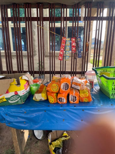 The image shows an outdoor market stall in Unguwar Sarki, Nigeria. A table draped with a blue plastic sheet displays various food items, including loaves of bread in orange packaging, a box of Cherie noodles, and a bag of eggs. Several small, colorful packets, likely condiments or snacks, are hung on a wire in the background. A green plastic basket sits on the table, and a transparent plastic container is visible next to it. The stall is set up against a weathered wooden fence and concrete structure, with windows and foliage visible in the background. The lighting suggests it is daytime.
