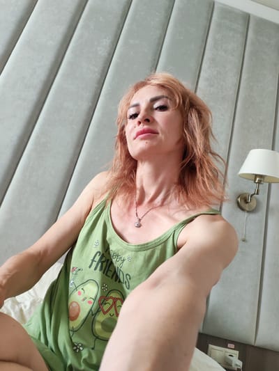 A woman with light brown hair and visible makeup is sitting on a bed with a light grey, vertically tufted headboard. She is wearing a green tank top with a graphic of smiling avocados and the text "happy FRIENDS" printed on it. A silver necklace with a small charm is visible around her neck. To the right of the woman, on the headboard, is a wall-mounted lamp with a white shade. A power outlet with a white plug is visible on the wall below the lamp. The scene is indoors, likely in a bedroom. There is no specific indication of this location being in Yoshkar-Ola, Russia.