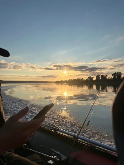 The image depicts a person's hand and arm in the foreground, with their finger extended towards the water. They are in a boat, with fishing rods visible. One fishing rod has a green lure attached. The boat is moving, creating a wake in the water. The scene is set on a body of water, likely a lake or river, with a tree-lined shore in the distance. The sun is setting, casting a warm glow on the water and clouds. The sky is a clear blue with some wispy clouds. The overall mood is peaceful and serene, suggesting a relaxing fishing trip at sunset in Ol'khovskoye, Russia. No visible text is present in the image.