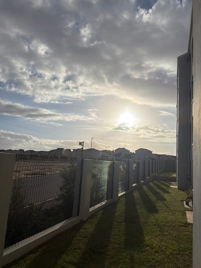 The image shows a view from what appears to be a high vantage point, likely a building. The sky dominates the scene with a mix of bright sun and clouds. Below the sky are buildings and a fence line running parallel to the buildings. Adjacent to the fence line is a green grassy area. The image is taken from what may be a balcony in Cape Town, South Africa. There are no people visible in the image. The architectural style of the buildings is modern. The presence of the fence suggests a boundary between the buildings and the grassy area. Shadows cast on the grass indicate a time of day when the sun is at an angle.