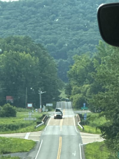 The image shows a two-lane road with yellow double lines, ascending a hill. Two vehicles are visible on the road, both appearing to be SUVs or similar passenger vehicles. The road is bordered by green grass and trees. In the background, a densely wooded hillside rises steeply. The sky is not visible. A black car mirror is partially visible in the upper right corner, suggesting the photo was taken from inside a vehicle. Several signs are visible along the side of the road, including a street light and what appear to be informational or directional signs. The overall scene is rural and hilly.