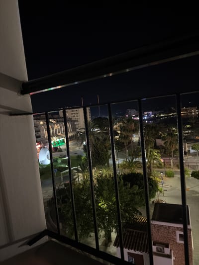 The image is a nighttime view from a balcony overlooking a street in Sant Antoni De Portmany, Spain. In the foreground, a black metal railing divides the view. Beyond the railing, a street is lined with buildings, palm trees, and lampposts. One building features an arched facade illuminated by warm lights. Across the street, a large white spherical object is visible. The sky is dark, with faint lights from distant structures and the sea visible on the horizon. The overall impression is that of a lively resort town at night.