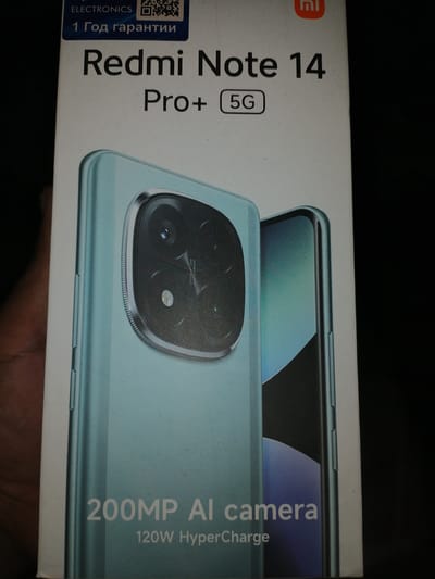 A product box for a "Redmi Note 14 Pro+ 5G" smartphone is presented against a dark background. The box is primarily white with some areas featuring a teal gradient, and it displays promotional text and imagery. Prominent text includes "Redmi Note 14 Pro+ 5G" and "200MP AI camera 120W HyperCharge." A graphical representation of the phone's rear camera system, featuring multiple lenses, is visible. At the top of the box, there is a "1 Год гарантии" (1 Year Warranty) label in Russian, along with an "ELECTRONICS" label and a QR code. The "mi" logo is also present in the upper right corner. There is no discernible connection to the city of Juma, Uzbekistan, in the provided image.
