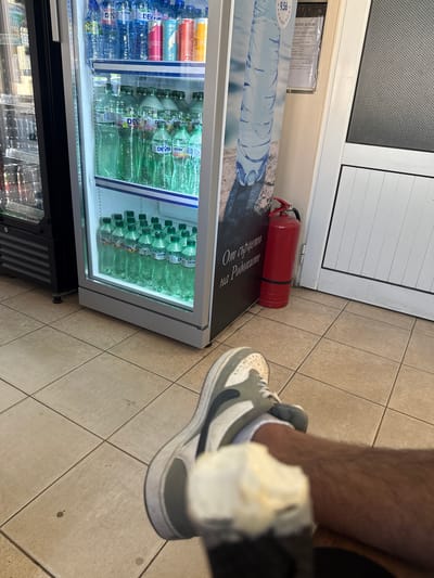 A low-angle, eye-level shot shows a person's legs and feet in the foreground, with a commercial refrigerator filled with bottled water and cans to the left. The refrigerator is illuminated from within, showcasing rows of plastic water bottles and various canned beverages. To the right of the refrigerator, a red fire extinguisher stands upright against a wall, and a door with a textured glass panel is visible. A portion of a wall poster with a large image of a water bottle is also visible on the left side of the frame. The floor is tiled with square, beige tiles. The overall setting appears to be an indoor establishment, possibly a store or a rest stop. The location in the city of Strazhitsa, Bulgaria is not explicitly indicated in the visual information provided.