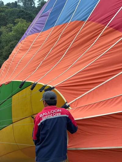 The media file captures a close-up view of a hot air balloon from the inside, with a person wearing a "BALLOON Ground Crew" jacket visible. The balloon's fabric is composed of large, colorful panels in shades of red, orange, blue, purple, yellow, and green, separated by white seams. The person, seen from behind, is wearing a gray baseball cap and a blue and red jacket with white text that reads "BALLOON" and "Ground Crew". They appear to be attending to the balloon's rigging, with dark fabric loops and suspension lines visible. The scene is set outdoors, with hints of green foliage and a cloudy sky visible in the upper left corner, suggesting it might be an overcast day. The overall impression is one of preparation for a hot air balloon flight, with the ground crew member engaged in their duties. The location context provided is Vang Vieng, Laos.