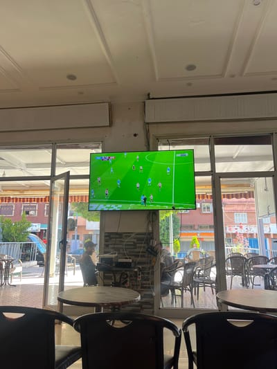 The interior of a cafe or restaurant in Ouaouizert, Morocco, is shown, where a flat-screen TV displays a soccer match in the middle of a bright day. A person with a dark beard is sitting in the foreground. The windows offer a view of the street and buildings of the city outside. There are wooden tables and chairs near the window. The interior walls are white and have ornate moldings.