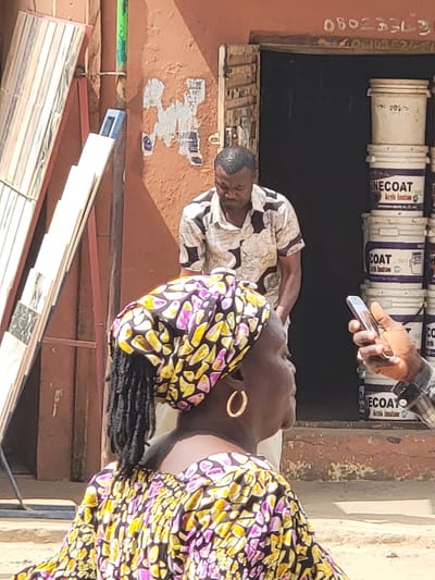 In Maiburiji, Nigeria, the image shows three individuals near a building displaying paint buckets labeled "ONE COAT." One man in a patterned shirt is seated. A second man holds a phone. A woman wearing a colorful headwrap and earrings stands nearby. Tiles lean against the building's exterior, which has a phone number painted on it.