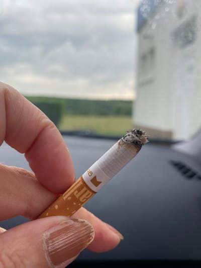 An extreme close-up shot shows a person's hand holding a lit cigarette. The cigarette is partially burned, with ash at the tip. The cigarette filter is white with an orange band and text that appears to be "ar" and "06". The background is blurred, showing a cloudy sky, green trees, and a white building. The overall impression is one of a casual moment outdoors. The location of Bílina, Czechia is not visually present in the image.