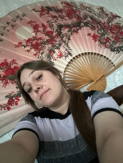 A selfie of a woman lying on a surface covered with a large, ornate Asian fan. The fan is decorated with red floral patterns and has a wooden frame. The woman has brown hair, light skin, and is wearing a gray and white striped top with black trim. A brown shoe is visible in the upper right corner of the image. The location is not evident from the image.