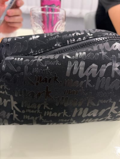 The image shows a black cosmetic bag with a repeating "Mark" logo pattern. The bag has a zipper closure and is placed on a white surface. In the background, a person's hand holding a smartphone is visible, along with a glass of liquid and a pink can of what appears to be a beverage. The scene is indoors, likely a table setting. The image does not contain any elements that would allow for the identification of the city of Novi Iskar, Bulgaria.