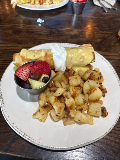 A plate of food sits on a wooden table. The plate contains a breakfast burrito covered in hollandaise sauce and a dollop of sour cream, a side of fried potatoes, and a metal cup filled with cut strawberries, pineapple, grapes, and blueberries. The plate has a textured, decorative edge. The photograph was taken in Bryan, Texas, United States.