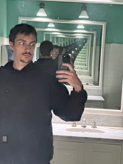 A person wearing a black hoodie is taking a selfie in a bathroom mirror. The mirror setup creates a "mirror effect," showing multiple reflections of the person and the surrounding bathroom. The bathroom has green walls and white trim. A sink with silver fixtures is visible, along with a white cabinet. The person's reflection extends into the distance, gradually diminishing in size.
