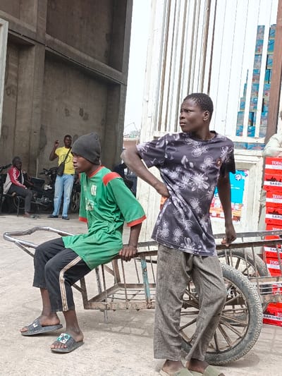 In Fagge C, Nigeria, an outdoor scene depicts five males and commercial activity under bright daylight. In the foreground, a young male with dark skin sits on a rusty, two-wheeled metal pushcart. He wears a dark grey beanie, a green t-shirt with red trim, black track pants, and black slide sandals, looking left. Standing to his right, another young male with dark skin wears a black t-shirt with white floral patterns, loose light brown pants, and light green slide sandals. He stands with his right hand on his hip, also looking left.
In the midground, to the left of the pushcart, a male in a yellow t-shirt and light blue jeans stands, gesturing with his raised right arm. Behind him, another male in a red and white jacket sits on a motorcycle. A second motorcycle is parked nearby. To the far right, a tall stack of red and white boxes, labeled "TODAY" and "SARDINE," stands beside a light-colored storefront or kiosk with various items and posters. Above these boxes, a vertical stack of blue and white boxes is visible against a tall, corrugated metal structure. The background features unpainted concrete walls of a building on the left and a bright, hazy sky. The ground is paved, appearing worn.