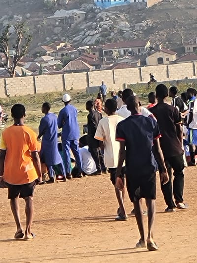 A group of approximately 15-20 people are lined up on a dirt field in Ibo, Nigeria. They appear to be performing some kind of exercise, as they are bending forward from the waist with arms extended. The people are wearing a variety of clothing, including different colors of shirts and pants, and some are wearing head coverings. A brick wall runs along one side of the field, and behind the wall are houses and other buildings. In the background, there is a hillside with rocks and vegetation.