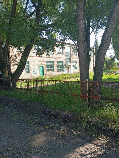 A two-story building with a white brick facade and green-trimmed windows stands behind a rusted metal fence. Trees with green foliage are in the foreground, partially obscuring the building. The ground in front of the fence is covered in gravel and dirt, with grass growing along the fence line. The building appears to be in a rural or suburban setting, possibly in the village of Novokopylovo, Russia, given its architectural style and the surrounding natural elements.