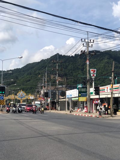 The image shows a street scene in Kamala, Thailand, with a focus on the intersection and the buildings lining it. A prominent blue and gold archway, likely a gateway or decorative structure, spans the road. Numerous utility poles with tangled wires crisscross the sky above, interspersed with lush green hills in the background.
On the right side of the street, a 7-Eleven convenience store with its distinctive red and green branding is visible, along with other shops, including a pharmacy with Thai and Cyrillic signage. A traffic light shows a green arrow for left turns.
Several people on motorcycles are present, some stopped at the intersection and others moving along the road. A red car is also visible among the vehicles. The road surface is asphalt, with a painted white line dividing the lanes. The sidewalks are lined with red and white striped curbs. The sky is partly cloudy.