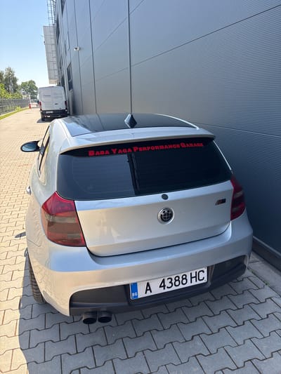 The image shows the rear of a silver BMW hatchback parked on a paved area outside a modern industrial building. The building has a grey corrugated metal exterior. A red sticker on the rear window of the car reads "BABA YAGA PERFORMANCE GARAGE". The car has a Bulgarian license plate with the registration "A 4388 HC". In the background, a white delivery van is parked, and trees and a fence are visible. The sky is clear and blue. The overall scene suggests a commercial or industrial location in Krivina, Bulgaria.