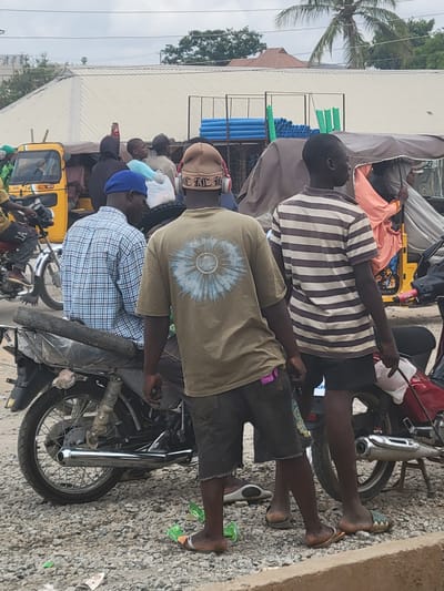 The image shows several people sitting on motorcycles in Suleja, Nigeria. The people are wearing casual clothing. One man wears a striped shirt, another a green shirt with a sun pattern on the back, and a third wears a blue plaid shirt. The motorcycles are parked on a street with gravel and trash. Other people and vehicles, including a yellow auto rickshaw, are visible in the background. Blue and green pipes are also being transported on one of the motorcycles.