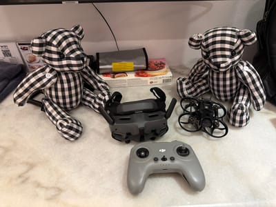 In Ban Phonsavang Neua, Laos, on a mottled white surface, are two black and white checkered teddy bears. Between them, a pair of FPV goggles and a drone controller are laid out. To the left of the goggles is a small black drone. Behind the drone and goggles are a gray fabric pouch with a yellow warning label and a box of pastries. A black backpack is visible on the far right.