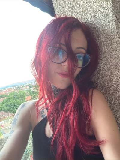 This is a vertical, candid, selfie-style photograph taken outdoors, likely during the day with bright lighting. The subject is a person with long, bright red hair, wearing glasses with light blue tinted lenses. Her face is partially obscured by her hair, but her lips, painted with pink lipstick, are visible. She appears to be lying down on a textured surface, possibly a carpet or stone. Her left arm is visible, resting near her head, and features several tattoos. She is wearing a black tank top.

In the background, out of focus, is a scenic view of a town with red-tiled roofs and lush green trees, suggesting a rural or semi-rural setting. The bright sky indicates it's daytime, and the lighting suggests clear weather. The overall mood seems relaxed and casual. No text is visible in the image. The location context of Svilengrad, Bulgaria, aligns with the visual cues of the town and vegetation.