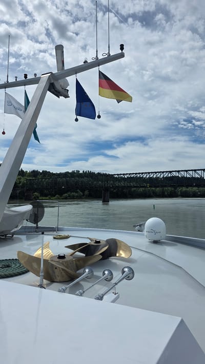 The image captures a detailed view from the deck of a boat navigating a wide river, identified as being in the city of Passau, Germany. In the immediate foreground, the clean white deck of the vessel is prominent, displaying two large, multi-bladed bronze ship propellers lying flat. Adjacent to them are three polished silver air horns, arranged in a line. A coil of green rope is visible to the left of the propellers, and a white, dome-shaped satellite antenna, branded "OCEAN SAT," is positioned further back on the deck.
Above the deck, a robust grey metallic mast structure rises, equipped with various navigation lights, antennas, and a white cylindrical radar dome. From this mast, four flags are hanging: a white flag with a blue logo (possibly "Dunay 24/7"), a teal-colored flag, a blue flag adorned with yellow stars (the European Union flag), and a black, red, and gold tricolor flag (the German national flag).
The background features a broad river with calm, grey-green water, extending towards a distant shore. A green navigational buoy is faintly visible in the water. Spanning the river is a dark, multi-span metal truss bridge, supported by sturdy concrete piers, with dense green forest lining the riverbanks behind it. The sky above is partly cloudy, with scattered white cumulus clouds against patches of blue, and a faint white contrail streaks across the upper right portion of the frame. No individuals are visible in the scene.