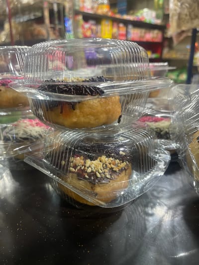 The image shows a stack of donuts in clear plastic containers, likely on a shelf in a store. The donuts are glazed and topped with various toppings, including chocolate sprinkles and chopped nuts. In the background, shelves filled with products are visible, suggesting a retail environment. The lighting is artificial, and the focus is on the donuts in the foreground. The scene appears to be in a store located in Barat Yaman, Indonesia, as indicated by the context provided.