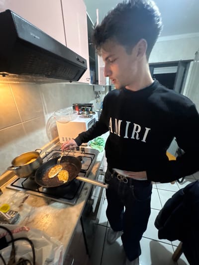This image depicts an indoor kitchen scene in the city of Bragança, Portugal. A young adult male is standing at a gas stovetop, actively engaged in cooking. He possesses a light skin tone and dark hair, and is dressed in a black long-sleeve sweatshirt featuring "AMIRI" in white lettering across the chest, dark blue jeans, and a dark belt with a gold-colored double-C shaped buckle. His white underwear is visible above the waistband of his jeans.
The male subject holds a silver-colored fork in his right hand, using it to manipulate two pieces of light-colored, possibly breaded, food within a black frying pan situated on the front-right burner of a four-burner gas stove. A silver pot with a lid handle is positioned on the rear-left burner, containing a yellowish substance.
The kitchen environment includes a light-colored countertop, likely marble or granite, where a white plastic bag and other items are visible on the lower left. A black range hood with controls is mounted above the stovetop. Above the range hood, pinkish-brown overhead cabinets are present. A tiled backsplash covers the wall behind the stove. To the right, a white microwave oven rests on the countertop, topped with a wire rack. A light green cloth is situated near the microwave. Artificial lighting illuminates the area, casting light on the cooking surface. A window or door with a dark exterior is visible in the background, suggesting evening hours.