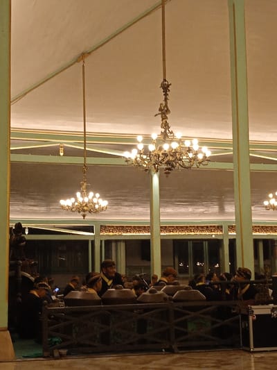 The image shows a large room with high ceilings and a light-colored tiled floor. Two ornate chandeliers with incandescent bulbs hang from the ceiling. There are several people sitting behind a wooden railing, wearing traditional clothing and hats. There is a large black speaker box sitting in the front corner of the room, near the railing. The scene is taken in a large building located at -7.559, 110.783 in Surakarta, Indonesia. This location is within the grounds of the Kraton Kasunanan Surakarta, the palace of the Surakarta Sunanate. The room appears to be a grand hall, perhaps a reception area or a performance space.