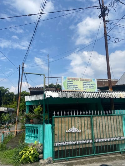 The image shows a storefront in Mojokerto, Indonesia, located at latitude -7.458 and longitude 112.446. The store, painted a vibrant turquoise, is situated on a narrow street and features a sign reading "SYAFIR GORDEN". The sign lists various products sold, including curtains, bed covers, bed sheets, and bedding. A green metal gate with intricate details separates the store from the street, and multiple power lines crisscross the sky. Lush greenery can be seen surrounding the store and on the opposite side of the street, while the sky is a bright blue with scattered white clouds.
