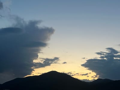 The image shows a silhouette of mountains against a twilight sky. A cross is visible atop the central peak. Dark clouds are present on the left and right sides of the frame, with patches of yellow and orange light from the setting sun peeking through. Two faint pink lights are visible in the upper right portion of the sky. The scene likely depicts the area around Sliven, Bulgaria, where the Karandila peak is a prominent landmark with a cross.