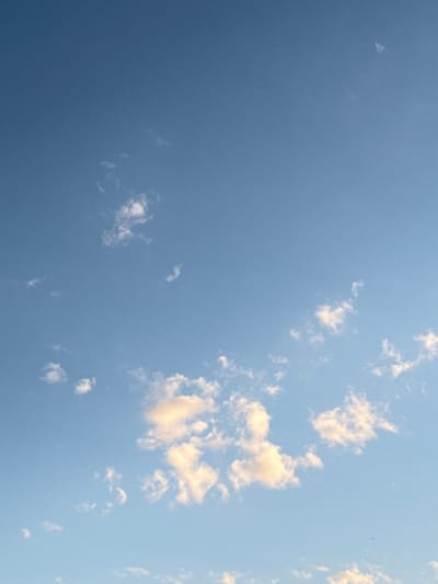 The image shows a blue sky with scattered clouds. Some of the clouds are tinted with a yellowish hue, likely from the sun. The location is assumed to be Прикамский, Russia, based on the prompt. The scene depicts a calm and sunny day with no visible people, subjects, objects, actions, or interactions other than the natural elements of sky and clouds.