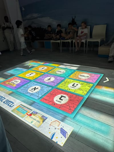 The image shows an interactive projection game on the floor of a room in Netanya, Israel. The game features a grid of colorful squares, each displaying Hebrew letters and mathematical symbols. Several children are visible in the background, watching the game. The room appears to be a classroom or activity space, with a mural on the wall depicting a starry night sky and silhouetted figures. Sunlight streams through a window, casting colorful patterns on the floor and the game.